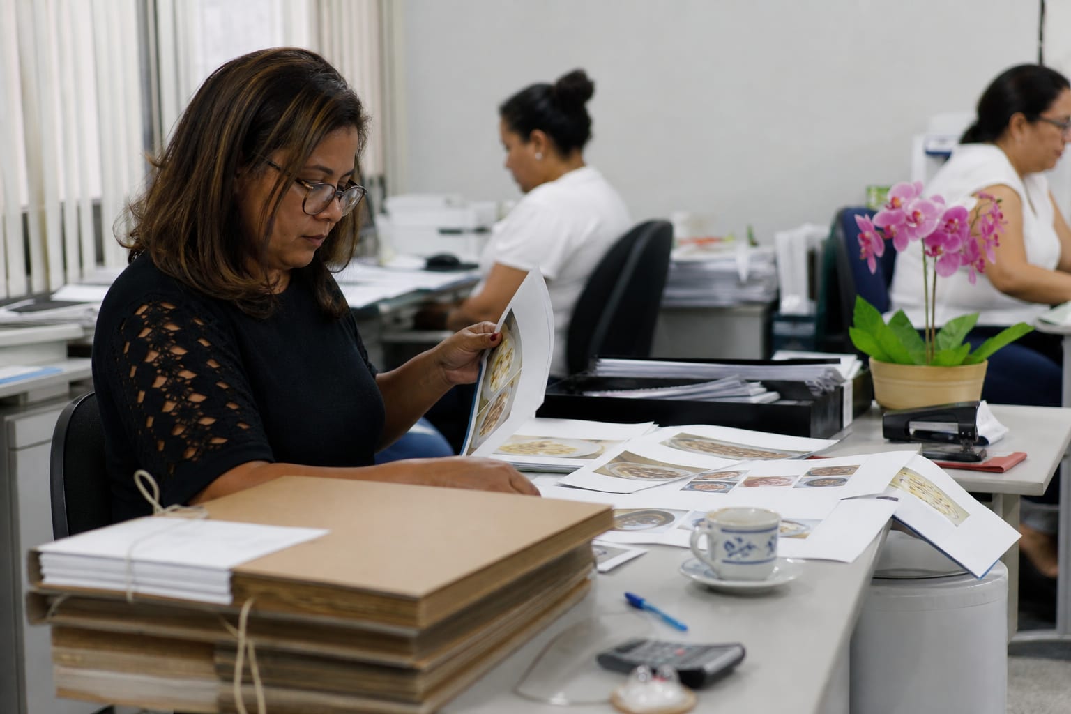 Mesa de secretaria municipal com pastas de arquivo, folhas impressas e calculadora durante a conferência de documentos
