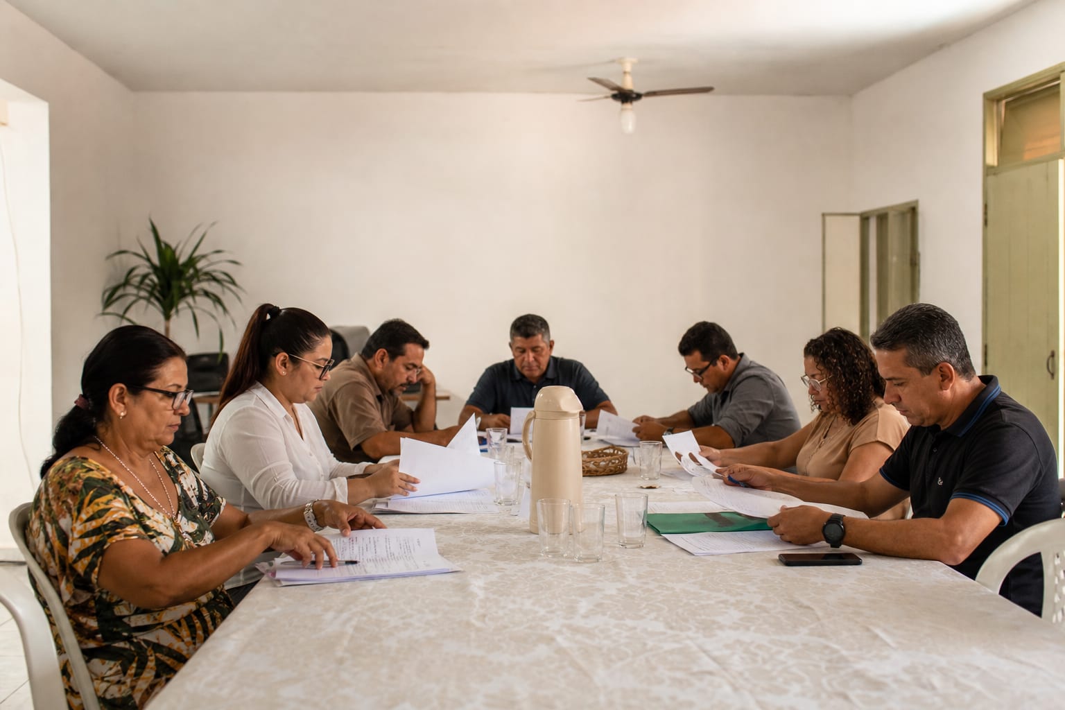 Reunião de conselho municipal com adultos folheando documentos impressos ao redor de uma mesa comprida