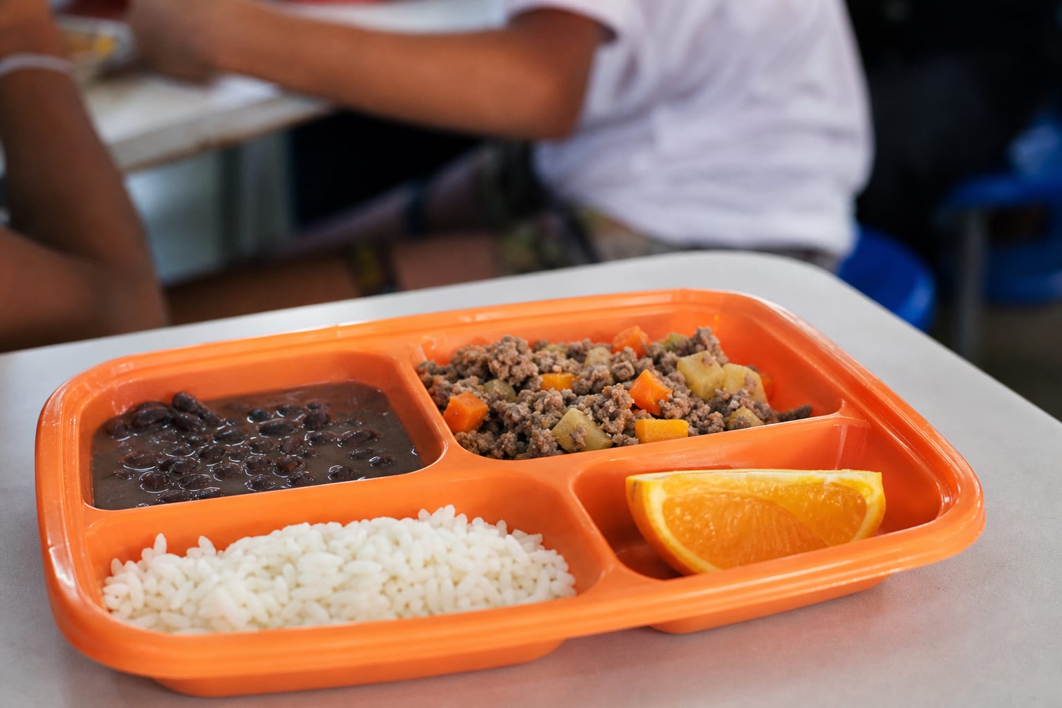 Bandeja laranja de refeitório escolar com arroz, feijão, carne moída com legumes e laranja cortada